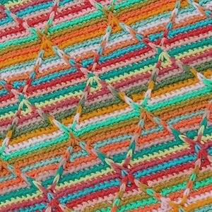 May include: Close-up of a colorful, hand-crocheted blanket. The blanket features a repeating pattern of diamond shapes, with a vibrant mix of red, orange, yellow, green, and blue yarn. The texture is visible, showcasing the craftsmanship.