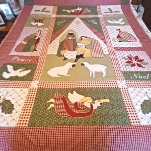 May include: A patchwork quilt featuring a central nativity scene with figures, sheep, and a star, surrounded by blocks depicting angels, doves, poinsettias, and holly. The quilt uses a color palette of red, green, and cream with gingham borders.