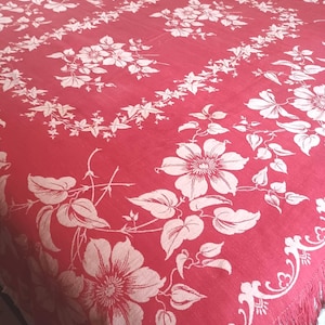 May include: A rectangular red bedspread featuring a repeating white floral and vine pattern. The fabric has a woven texture with a decorative border and a fringe edge along the bottom. The design includes large blossoms and leafy stems.