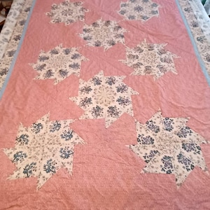 May include: A handmade quilt featuring a pink background with large, stylized floral starburst patterns in white and blue. The quilt has a light blue border and is likely queen or king size.
