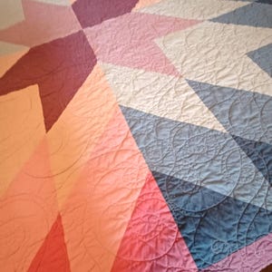 May include: A quilt featuring a geometric star pattern composed of diamond-shaped fabric pieces in shades of pink, purple, and blue. The surface displays intricate, continuous swirl quilting stitches throughout the fabric, creating a textured finish.