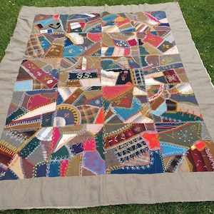 May include: A colorful patchwork quilt with a variety of fabric pieces in shades of blue, red, brown, and green. The quilt has a gray border and is laid out on green grass. The quilt is made of many different shapes and sizes of fabric.