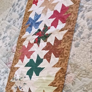 May include: A handmade table runner featuring a repeating pattern of white and colorful starburst shapes. The starbursts are in shades of red, blue, green, and brown, set against a brown patterned border. The runner is placed on a floral patterned bedspread.
