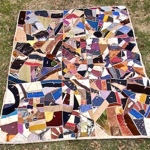 May include: A patchwork quilt with a variety of fabric pieces in shades of brown, blue, purple, and cream. The quilt has a geometric design with a white border and is laid out on a grassy surface.