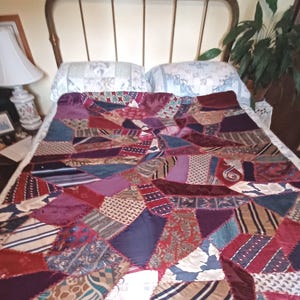 May include: A patchwork quilt constructed from various neckties in shades of burgundy, navy blue, purple, and gold. The quilt features an irregular, geometric arrangement of silk-like fabric pieces with diverse patterns, including stripes, paisley, and floral motifs, joined together with visible decorative stitching.