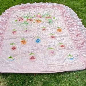 May include: A light pink bedspread with a ruffled edge. The bedspread features embroidered floral designs in shades of pink, orange, blue, and green. The bedspread is lying on a green grassy surface.