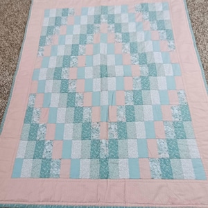 May include: A handmade quilt featuring a patchwork design in shades of light blue, pink, and white. The quilt has a rectangular shape with a pink border. The pattern includes squares and vertical stripes, creating a visually appealing textile.