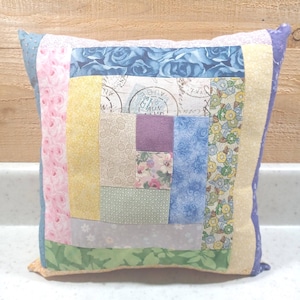 May include: A square throw pillow featuring a patchwork log cabin design. The fabric squares include patterns of blue roses, yellow textures, floral prints, and vintage-style postal stamps with circular postmarks in shades of blue, pink, and green.