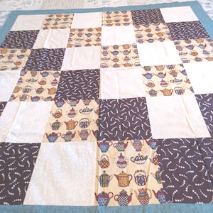 May include: A patchwork quilt featuring a repeating pattern of square blocks. The design alternates between solid white squares, dark brown squares with white curved dashes, and beige squares printed with colorful teapots. A teal border frames the quilt.