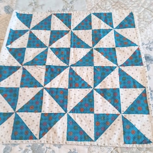 May include: A square quilt featuring a pinwheel pattern composed of alternating triangles. The design consists of solid white fabric with small black dots and a vibrant teal fabric patterned with small orange, pink, and green floral motifs.