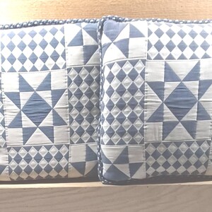 May include: Two square throw pillows with a patchwork design. The pillows feature a pattern of blue and white geometric shapes, including stars and diamonds. The edges are trimmed with a dark blue border.