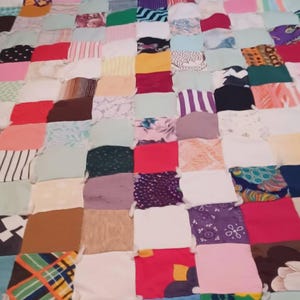 May include: A handmade patchwork quilt composed of numerous fabric squares in various colors and patterns. The quilt features a mix of solid colors, stripes, floral prints, and other designs, creating a vibrant and textured appearance. The squares are sewn together, forming a large, rectangular blanket.