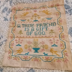 May include: A vintage cross-stitch sampler with the text "A TRUE FRIEND IS A GIFT OF GOD." The design features teal, yellow, and pink floral and bird motifs, framed by a teal border on a beige fabric.