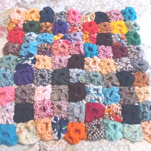 May include: A collection of numerous fabric hair scrunchies arranged in a grid pattern. The scrunchies feature a wide variety of solid colors including red, blue, yellow, orange, pink, and black, alongside various floral, plaid, and animal print patterns.