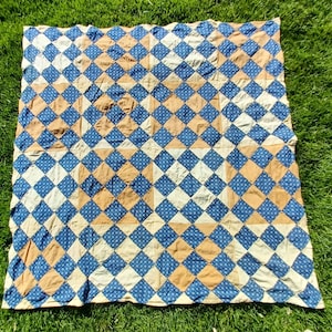 May include: A handmade quilt with a repeating diamond pattern in shades of blue, tan, and cream. The quilt is made of fabric squares sewn together, creating a geometric design. The quilt is lying on green grass.
