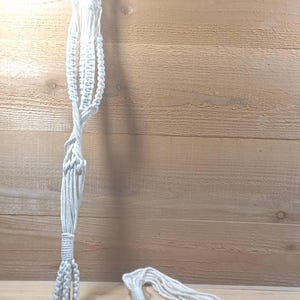 May include: A white macrame plant hanger featuring intricate knotted cord patterns, a sturdy metal ring at the top for suspension, and long, loose fringe tassels at the bottom, crafted from durable cotton rope.