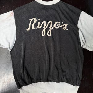 May include: A short-sleeve shirt with a dark charcoal gray body and light gray sleeves. The shirt features a ribbed light gray crew neckline and a wide ribbed light gray hem. The word Rizzo's is embroidered in a light cream script font on the back.
