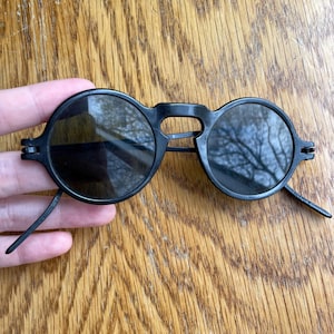 May include: Black round sunglasses with a bridge and thin arms. The lenses are dark, and the frame is a matte black. The sunglasses are held against a wooden surface.