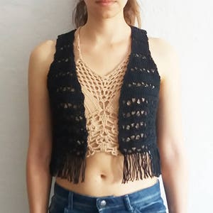 Puede incluir: Un chaleco de crochet negro con detalles de flecos. El chaleco es sin mangas y tiene un escote en V. Se lleva sobre un top de crochet beige.