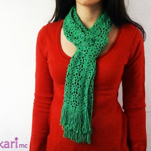 Op de afbeelding: Een groene gehaakte sjaal met franjes. De sjaal wordt gedragen door iemand die een rood shirt draagt.