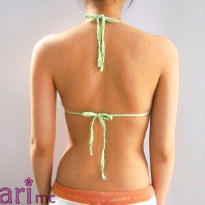 K&ouml;nnte beinhalten: Ein hellgr&uuml;nes, handgefertigtes H&auml;kel-Neckholder-Top. Das Top wird im Nacken und am R&uuml;cken gebunden und wird von hinten gezeigt. Das Top hat eine einfarbige hellgr&uuml;ne Farbe. Der untere Teil des Bildes zeigt den Rand einer orange-wei&szlig; gestreiften Shorts.