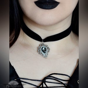 May include: A black velvet choker with a silver pendant featuring a large, faceted, dark gray gemstone. The pendant has a detailed, ornate design.