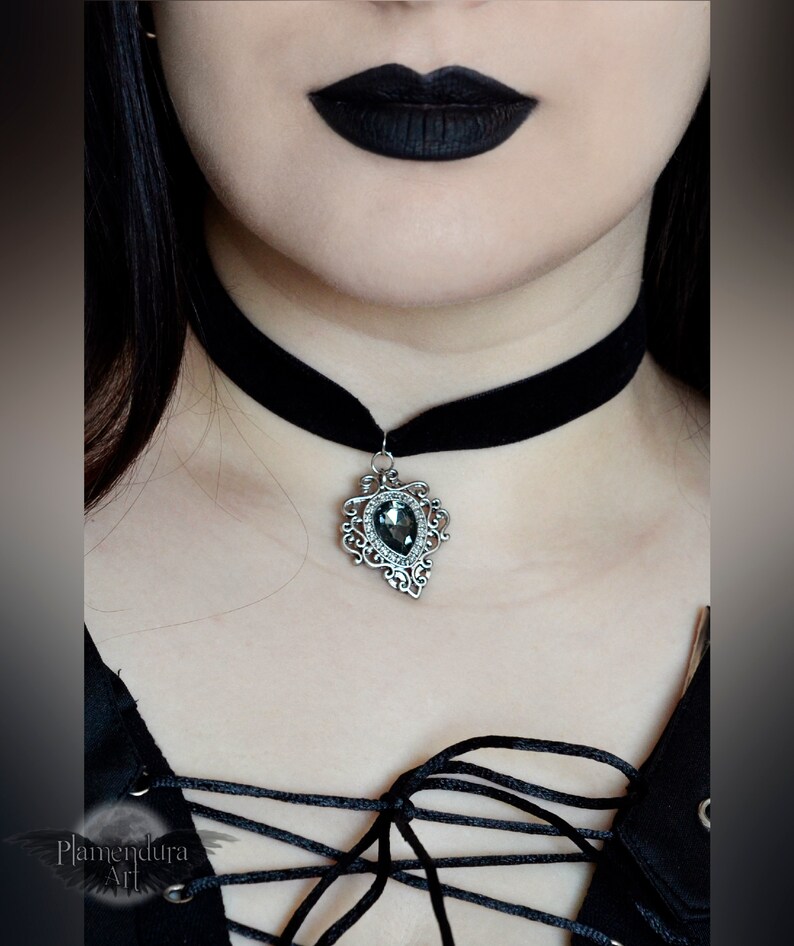 May include: A black velvet choker with a silver pendant featuring a large, faceted, dark gray gemstone. The pendant has a detailed, ornate design.