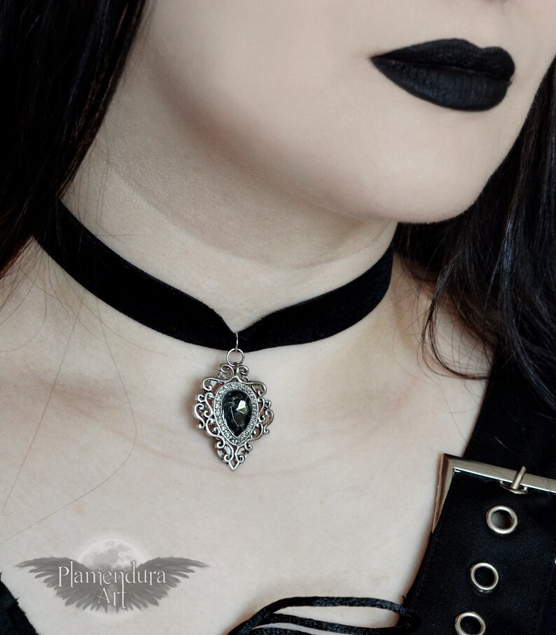 May include: A black velvet choker with a silver pendant featuring a large, dark gray gemstone. The pendant has a detailed, ornate design.