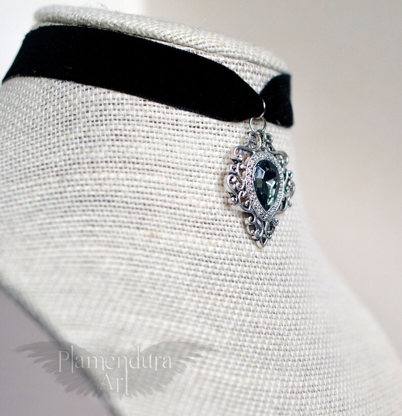 May include: A black velvet choker with a silver pendant featuring a large, faceted, dark gray gemstone in a heart-shaped setting.