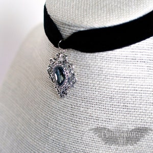 May include: A black velvet choker with a silver pendant featuring a large, dark blue gemstone. The pendant has an ornate, filigree design.