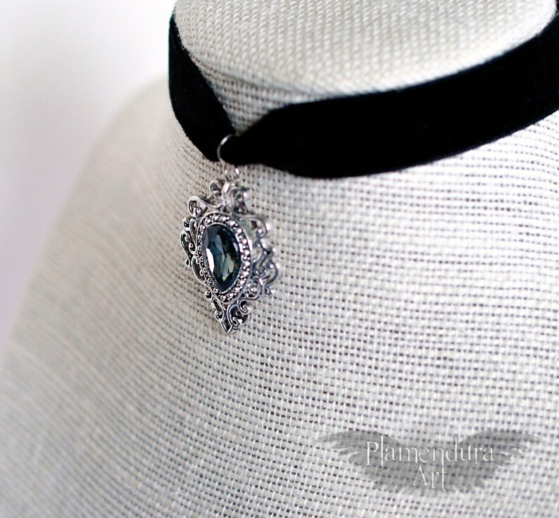 May include: A black velvet choker with a silver pendant featuring a large, dark blue gemstone. The pendant has an ornate, filigree design.