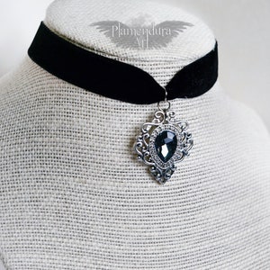 May include: A black velvet choker necklace with a silver pendant featuring a large, dark gray gemstone. The pendant has a detailed, ornate design.