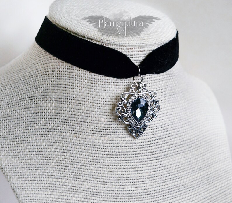 May include: A black velvet choker necklace with a silver pendant featuring a large, dark gray gemstone. The pendant has a detailed, ornate design.