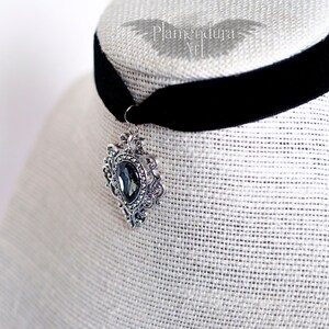 May include: A black velvet choker with a silver pendant featuring a large, dark gray gemstone. The pendant has an ornate, filigree design.