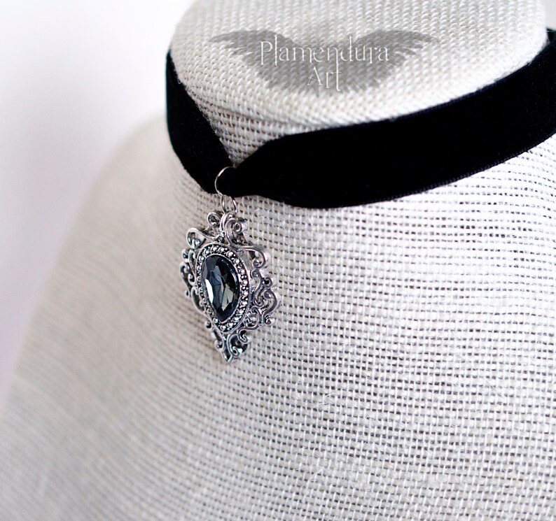 May include: A black velvet choker with a silver pendant featuring a large, dark gray gemstone. The pendant has an ornate, filigree design.