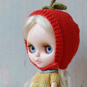 May include: A doll wearing a red knitted hat shaped like an apple with green leaves. The doll has blonde hair and blue eyes. The hat has a button on the side.