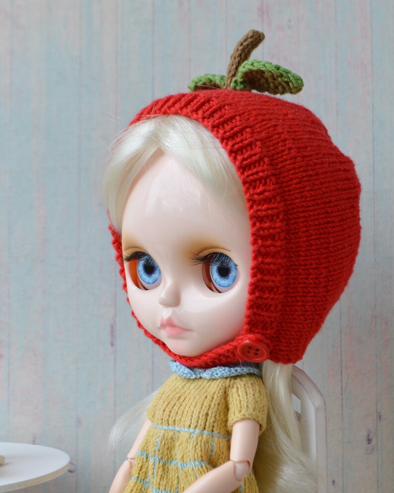 May include: A doll wearing a red knitted hat shaped like an apple with green leaves. The doll has blonde hair and blue eyes. The hat has a button on the side.