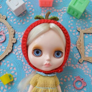 May include: A doll with blonde hair wearing a red knitted hat shaped like an apple with green leaves. The doll has blue eyes and is wearing a yellow knitted sweater.