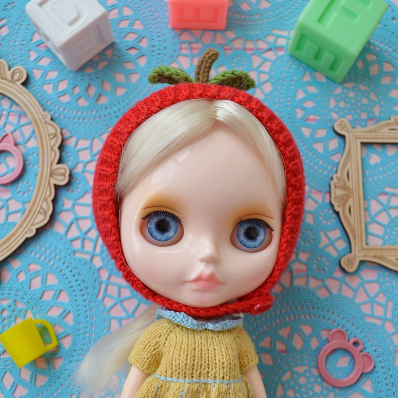 May include: A doll with blonde hair wearing a red knitted hat shaped like an apple with green leaves. The doll has blue eyes and is wearing a yellow knitted sweater.
