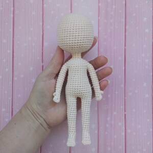 May include: A crocheted doll with a white body and no facial features. The doll is being held by a hand against a pink background with white stars.
