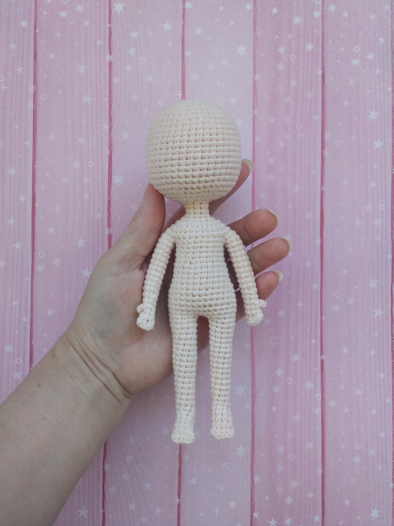 May include: A crocheted doll with a white body and no facial features. The doll is being held by a hand against a pink background with white stars.