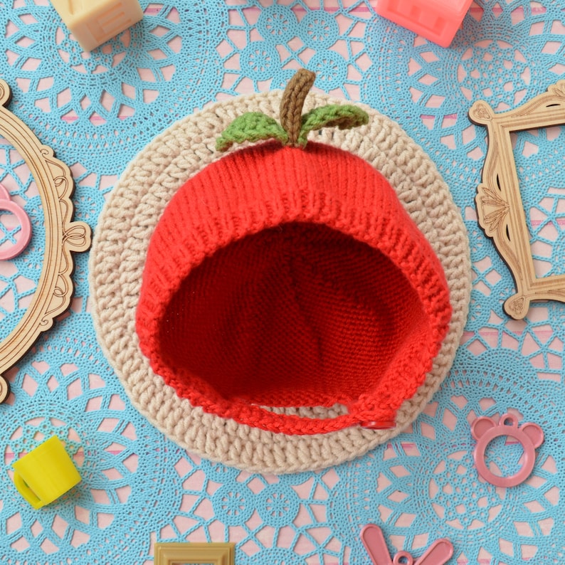May include: A red knitted baby hat shaped like an apple with a green leaf on top. The hat is on a crocheted beige background.