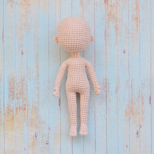 May include: A crocheted doll body in a light beige color against a blue wood background.