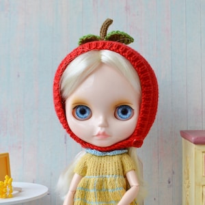May include: A doll with blonde hair wearing a red knitted hat shaped like an apple with green leaves. The doll has blue eyes and is wearing a yellow and blue checked dress.