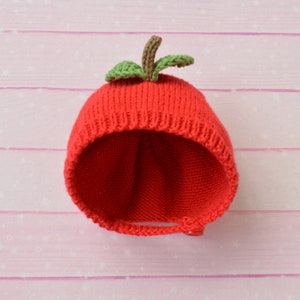 May include: Red knitted baby hat with a green apple stem and leaves on top. The hat is turned inside out and is lying on a pink wooden surface.