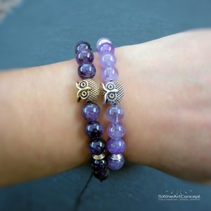 Puede incluir: Dos pulseras de piedra de amatista con dijes de búho. Una pulsera tiene un dije de búho dorado y la otra tiene un dije de búho plateado. Las pulseras están en la muñeca de una persona.