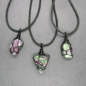 Peut inclure: Trois colliers avec des pendentifs en pierres précieuses vertes et roses enveloppés de fil noir. Les colliers sont sur un fond gris.