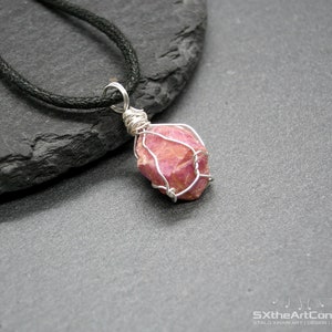 May include: A pink gemstone pendant wrapped in silver wire, hanging from a black cord necklace.