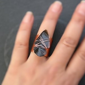 Op de afbeelding: Een koperen ring met een zwarte obsidiaan steen die is gewikkeld in koperdraad. De steen heeft de vorm van een traan en een ruwe, natuurlijke textuur.