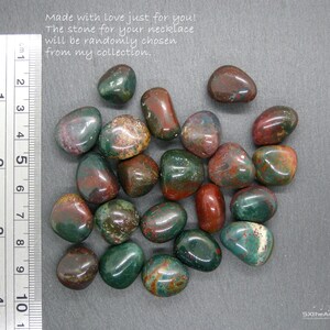 Op de afbeelding: Een verzameling gepolijste edelstenen in groen, rood en bruin. De stenen zijn glad en afgerond, met een liniaal die centimeters aangeeft. Tekst: "Made with love just for you! The stone for your necklace will be randomly chosen from my collection."